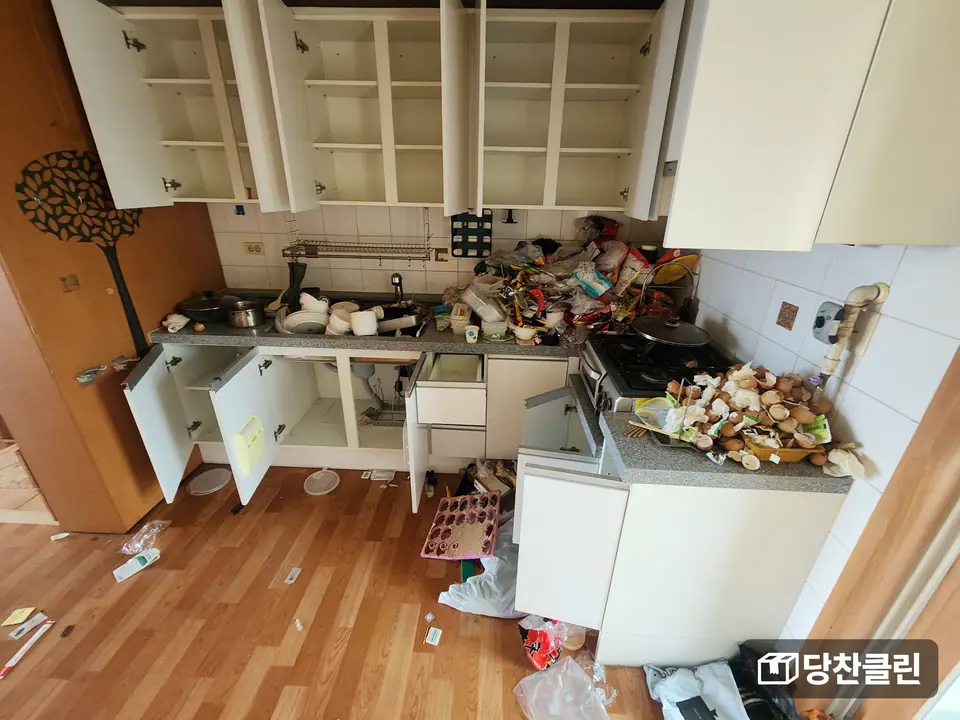 찬장 문이 모두 열려 있고 싱크대와 가스레인지 위에 쓰레기와 그릇이 쌓인 주방